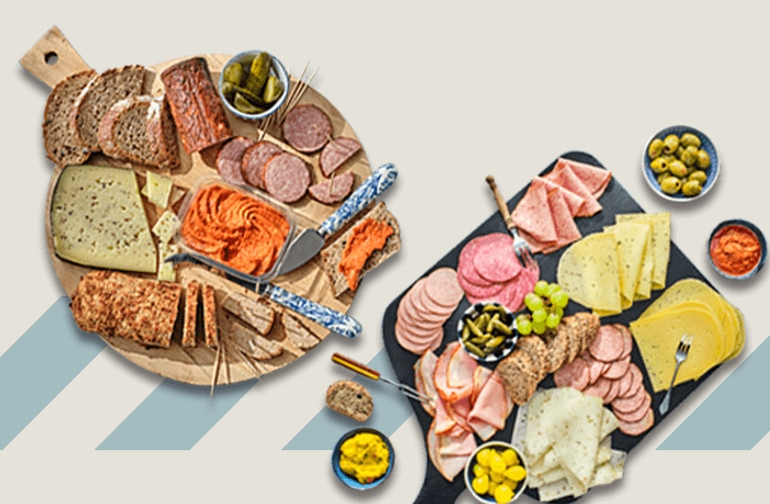Twee serveerplanken met een assortiment van vleeswaren, kazen, brood en augurken, omringd door schaaltjes met olijven en een rode saus.