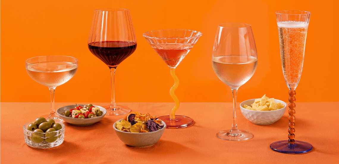 Verschillende glazen met wijn, cocktail en champagne, geserveerd met schaaltjes olijven, groentehapjes, chips en kaasblokjes tegen een oranje achtergrond.