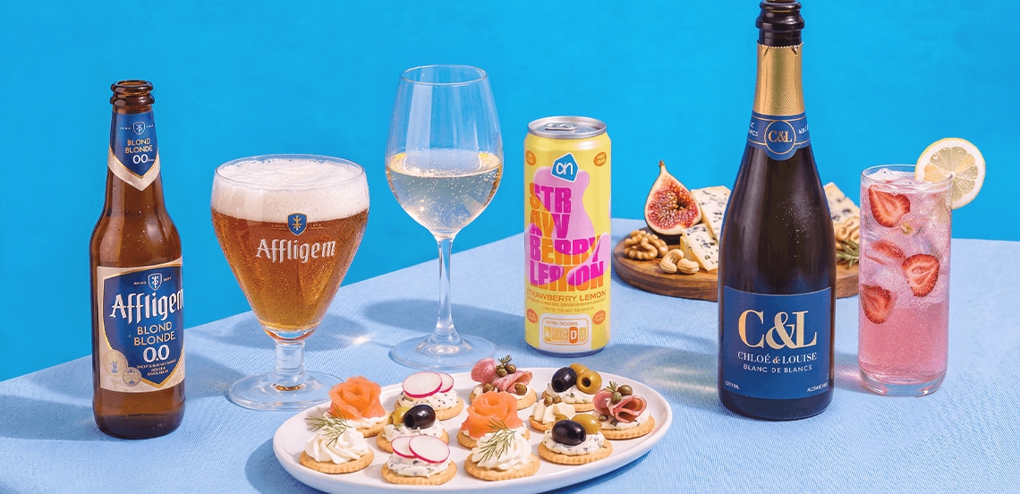 Assortiment van feestelijke hapjes, alcoholvrije Affligem blond bier, AH strawberry lemon frisdrank, een glas witte wijn, Chloé & Louise mousserende wijn, kaasplank met vijg en noten, en een verfrissende cocktail met aardbei en citroen op een lichtblauwe ondergrond.