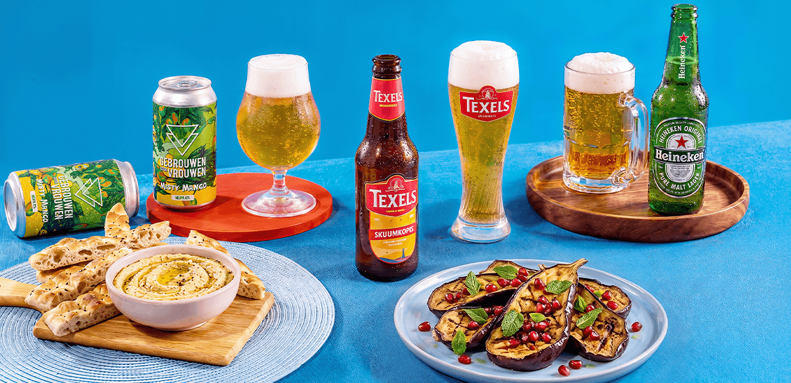 Afbeelding van verschillende biermerken waaronder Texels en Heineken, geserveerd in glazen en flessen, samen met een bord gegrilde aubergine met granaatappelpitten en verse kruiden, en een schaaltje hummus met flatbread, op een blauwe achtergrond.