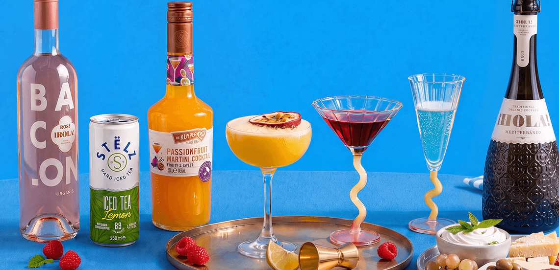 Assortiment van feestelijke drankjes en cocktails, waaronder BACON rosé, STËLZ hard iced tea lemon, De Kuyper Passionfruit Martini Cocktail en HO!LA Mediterráneo Brut, geserveerd op een gouden tray met verse frambozen, olijven, kaas en dip tegen een blauwe achtergrond.
