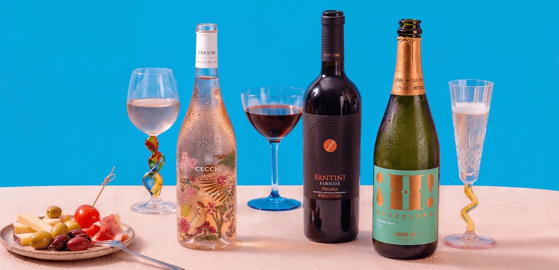 Drie flessen wijn (Cecchi rosé, Fantini rode wijn en Barcelona mousserende wijn) met bijpassende glazen, en een bord met kaas, olijven, tomaat en ham op een lichte tafel tegen een blauwe achtergrond.