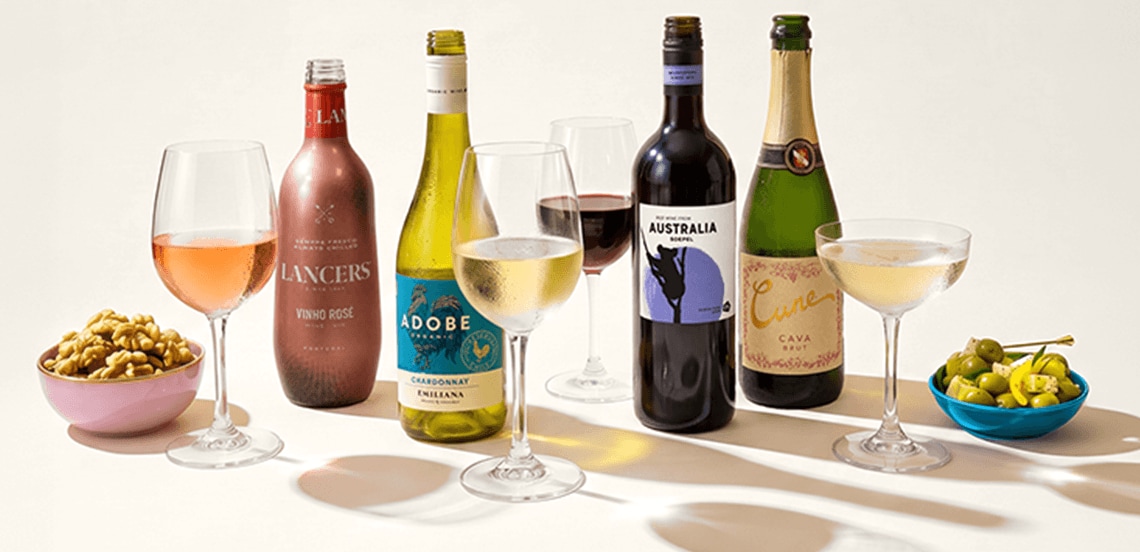 Vier flessen wijn (Lancers Vinho Rosé, Adobe Chardonnay, Australia Shiraz en Cune Cava Brut) met bijpassende glazen wijn, gepresenteerd met een schaaltje walnoten en een schaaltje groene olijven.