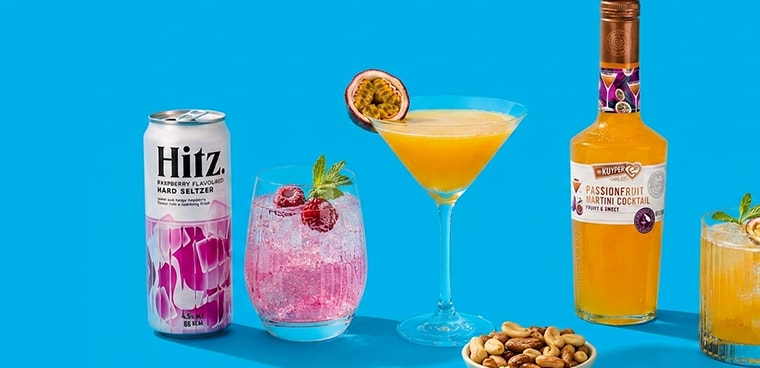 Blik Hitz Raspberry Hard Seltzer, glas met roze cocktail en frambozen, martiniglas met passievruchtcocktail van De Kuyper, halve passievrucht, en schaaltje gemengde noten op een blauwe achtergrond.