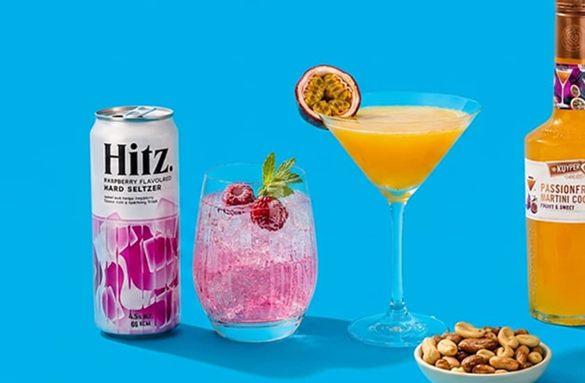 Blik Hitz Raspberry Hard Seltzer, glas met roze cocktail en frambozen, martiniglas met passievruchtcocktail van De Kuyper, halve passievrucht, en schaaltje gemengde noten op een blauwe achtergrond.