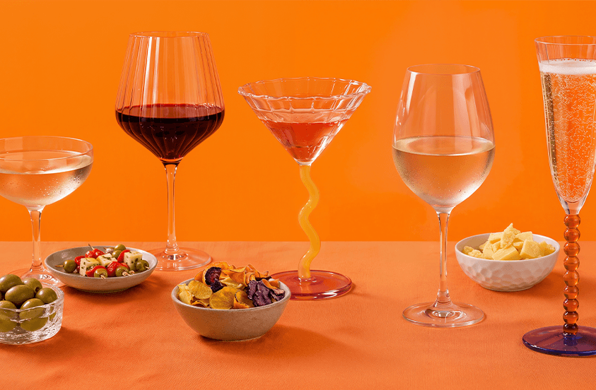 Verschillende soorten drankjes in elegante glazen met bijpassende borrelhapjes zoals olijven, kaasblokjes, groentechips en gemarineerde groenten, gepresenteerd op een oranje achtergrond.