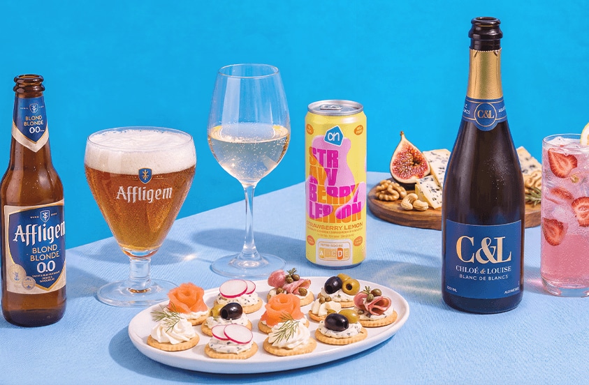 Een assortiment van feestelijke dranken en hapjes op een tafellaken, waaronder Affligem blond 0.0% bier, C&L Chloé & Louise Blanc de Blancs mousserende wijn, AH Strawberry Lemon drank, en een glas gevuld met aardbeien. Op de voorgrond staat een bord met luxe hapjes zoals crackers met zalm, radijs en olijven. Op de achtergrond liggen een vijg, blauwe kaas en walnoten.