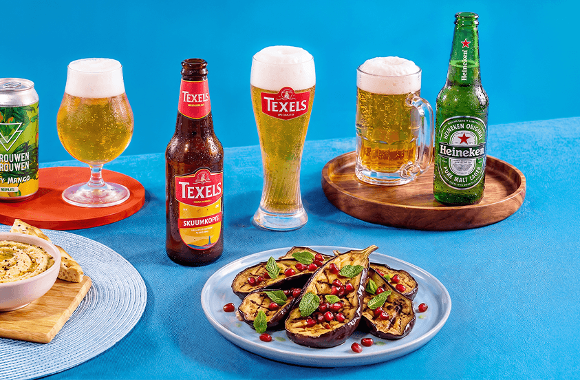 Afbeelding van verschillende biermerken waaronder Texels en Heineken, geserveerd in glazen en flessen, samen met een bord gegrilde aubergine met granaatappelpitten en verse kruiden, en een schaaltje hummus met flatbread, op een blauwe achtergrond.