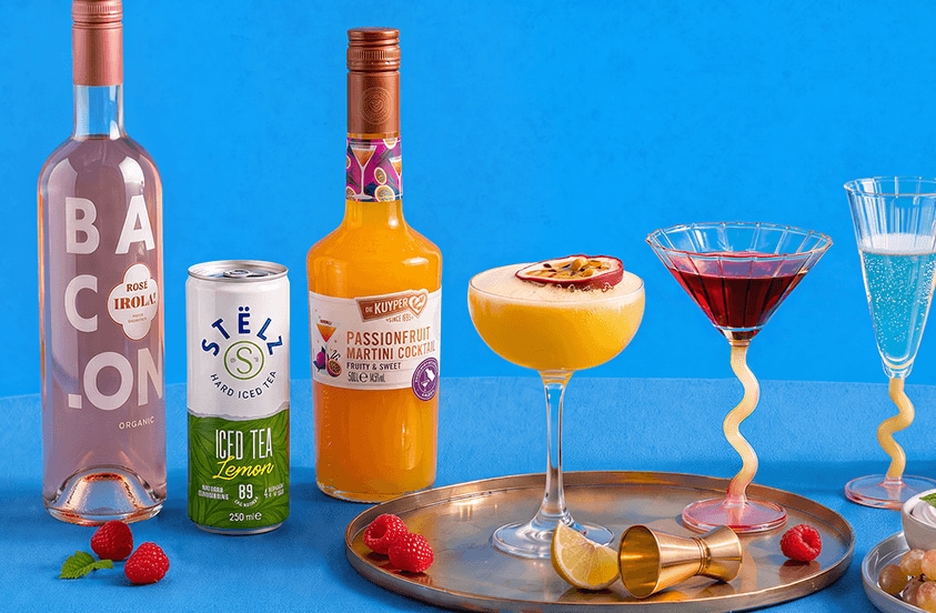Flessen BACON rosé, De Kuyper Passionfruit Martini Cocktail en een blikje STËLZ Iced Tea Lemon samen met verschillende cocktails in glazen, gepresenteerd op een goudkleurig dienblad met vers fruit als garnering, tegen een blauwe achtergrond.