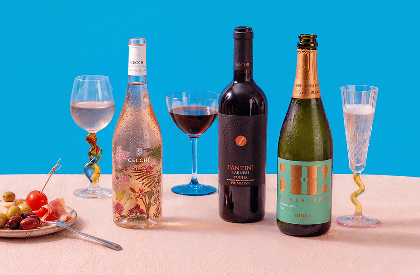 Drie flessen wijn, waaronder Cecchi rosé, Fantini Farnese rode wijn en Hola! Barcelona mousserende wijn, met bijpassende glazen en een bord met olijven, tomaat en prosciutto op een lichte tafel tegen een blauwe achtergrond.