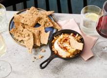 Ricotta als centerpiece van de gezonde borrelplank. Bak 'm in de oven en serveer met zelfgemaakte crackers.